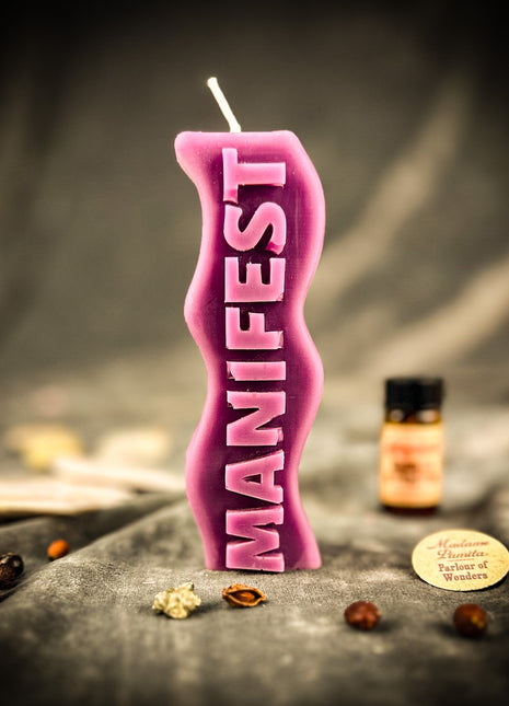 Pink candle with 'MANIFEST' text on a dark surface with decorative items.