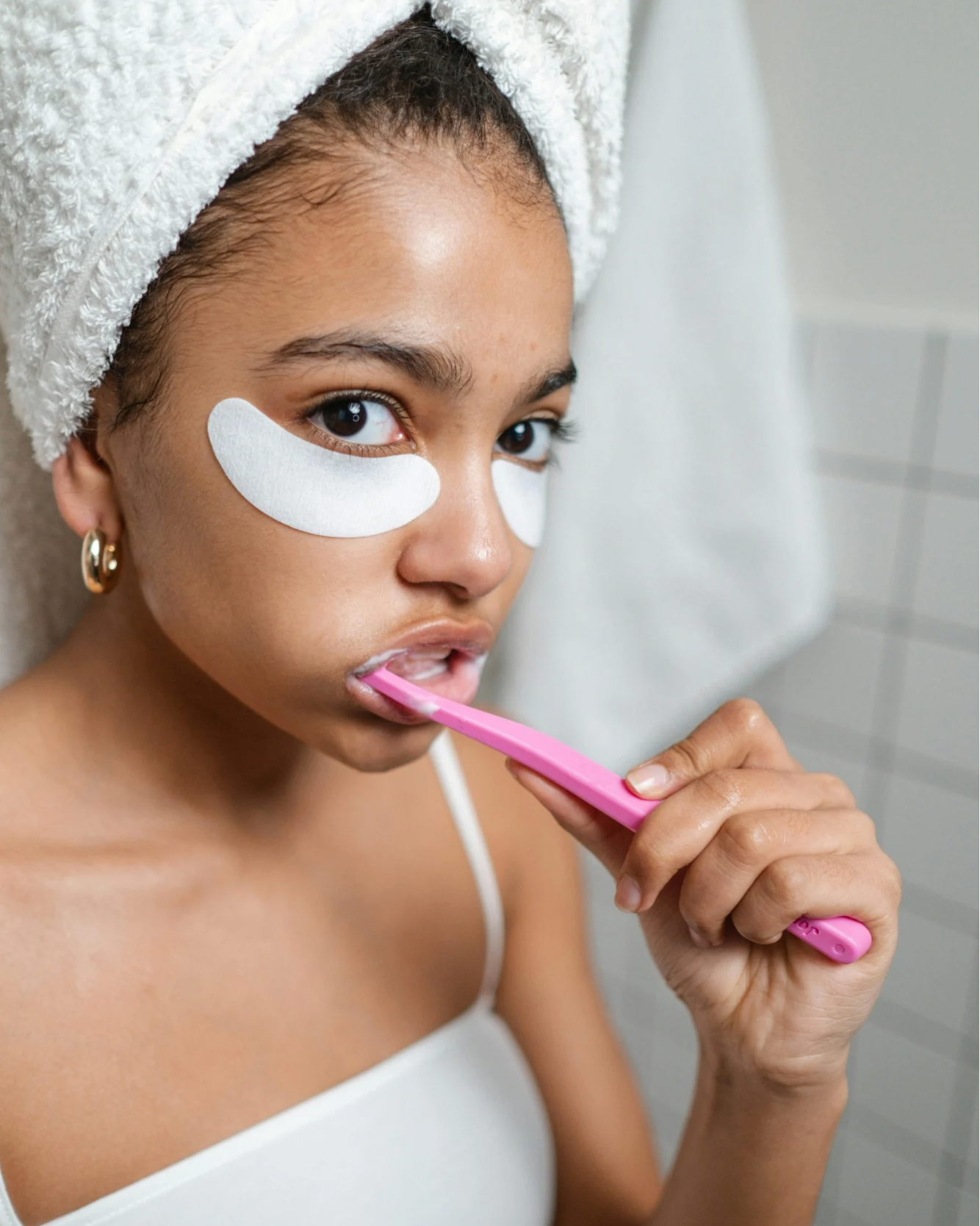 Woman brushing her teeth with a pink toothbrush, wearing white eye masks and a towel on her head.